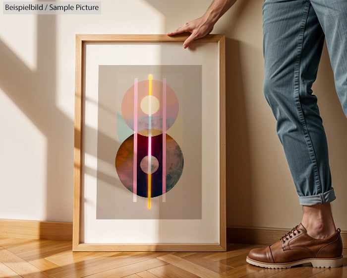 Person in jeans and brown shoes holding an abstract framed artwork on a wooden floor.