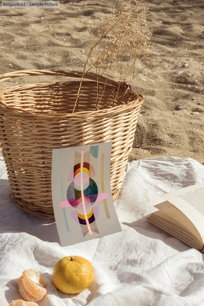 Beach scene with wicker basket, dried plants, abstract art, peeled citrus, and open book on a white cloth.