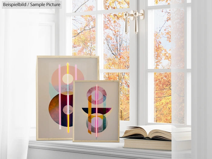 Colorful geometric artwork displayed on a windowsill beside an open book, with autumn trees visible outside.