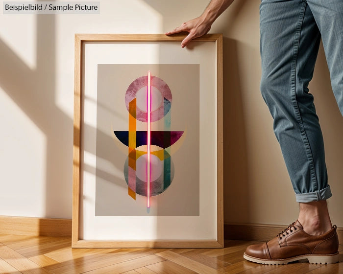 Framed geometric abstract art leaned against wall with person reaching toward it, wearing jeans and brown shoes.
