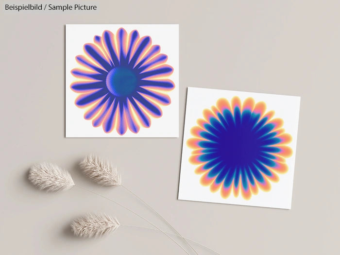 Two colorful abstract flower designs on white cards placed on a beige surface, with dried grasses beside them.