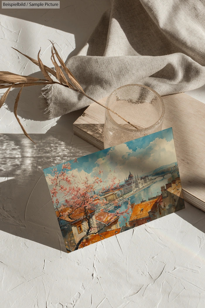 Postcard of a cityscape with cherry blossoms, positioned on a textured surface with a glass and dried plant.