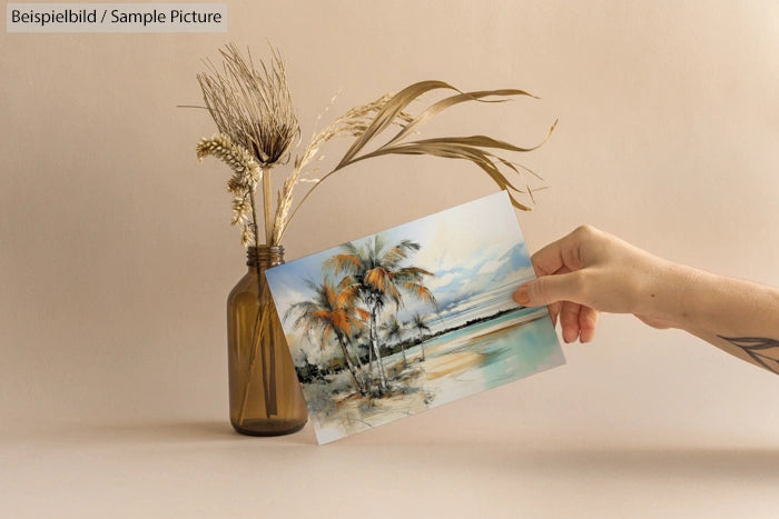 Hand holding beach painting in front of brown vase with dried grasses on neutral background.