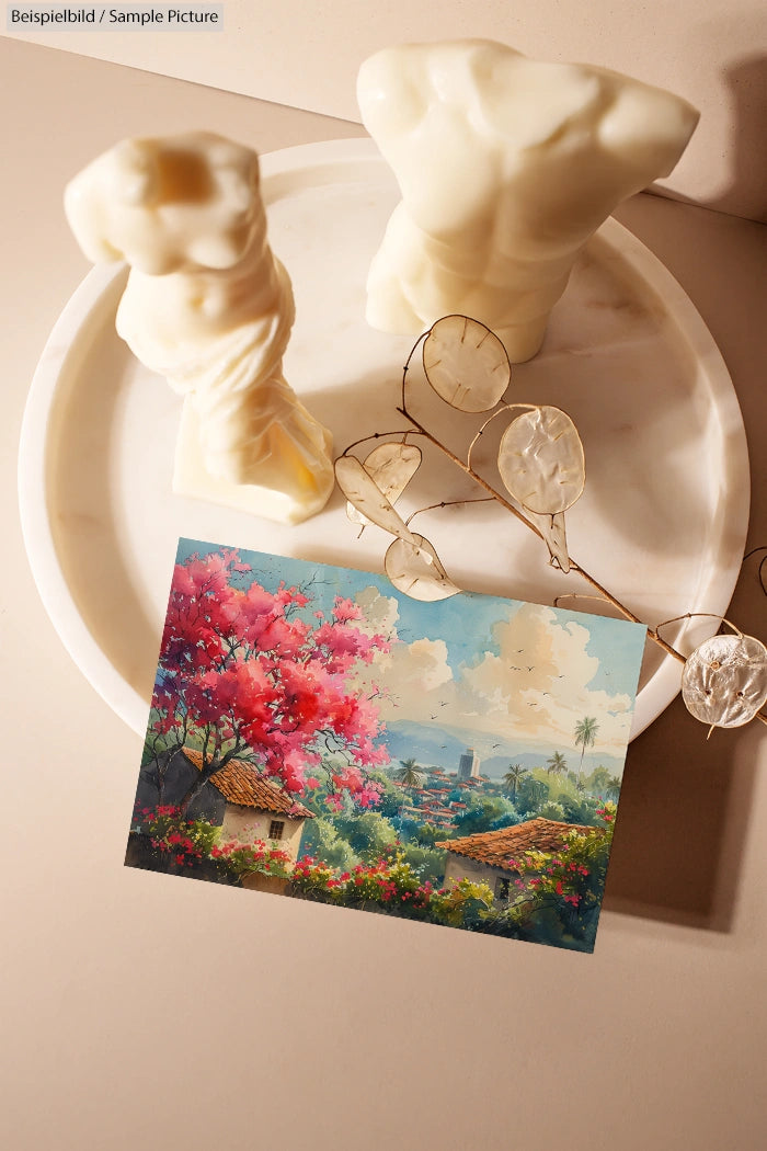 Postcard with vibrant landscape and city view next to sculpted candles on a round marble tray.