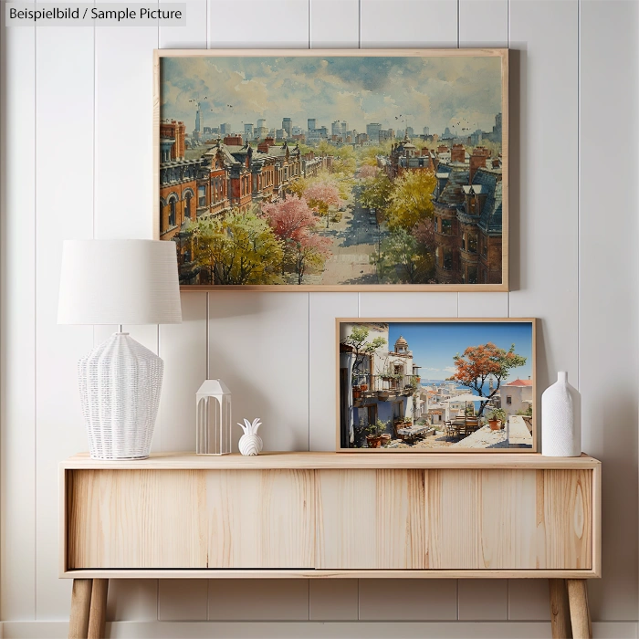 Two framed paintings on a wooden console; cityscape above, Mediterranean village scene below, against white paneled wall.