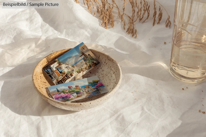 Ceramic bowl with postcards on white fabric, next to a glass of water and dried grass.