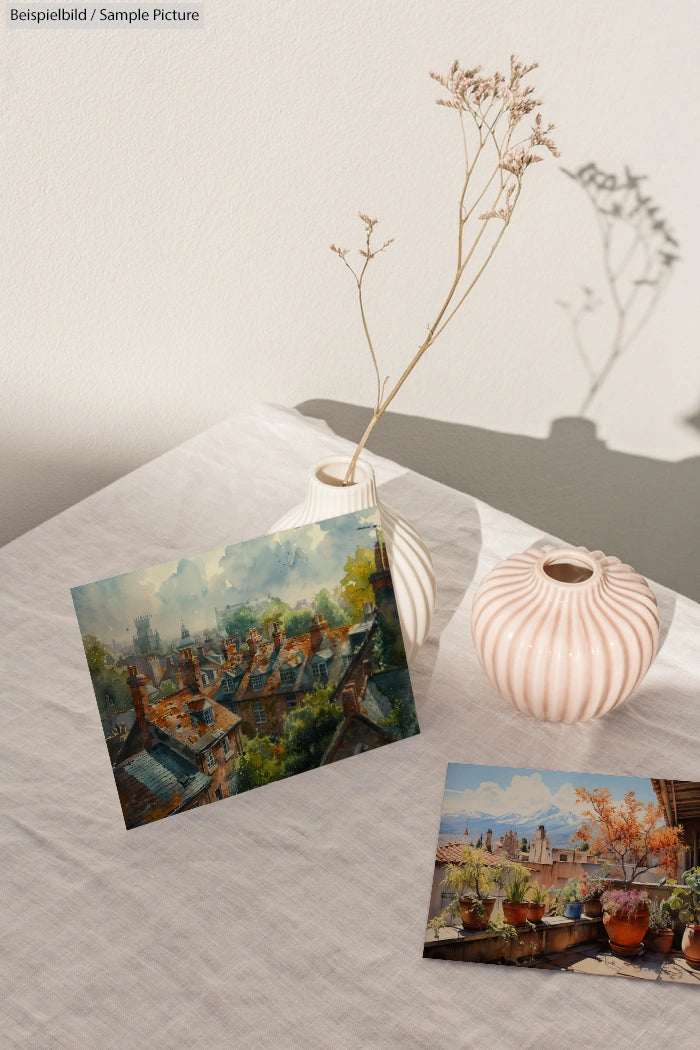 Two postcards on a table with a vase, displaying scenic landscapes and rustic rooftops, against a sunlight-filtered wall.