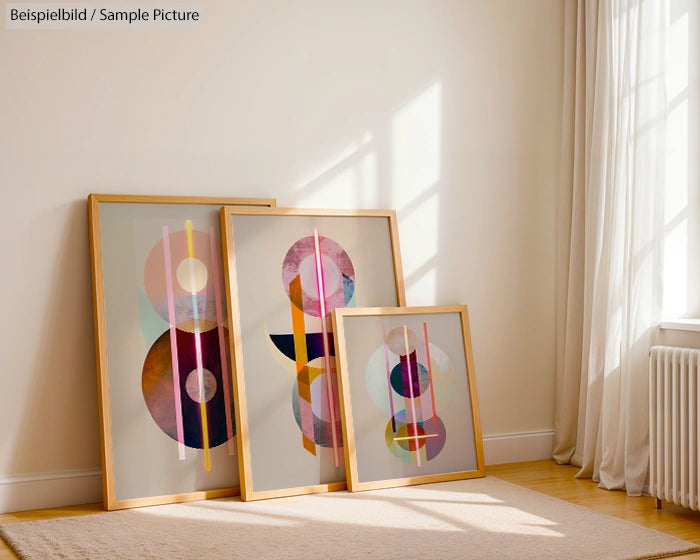 Three framed abstract artworks with geometric shapes and pastel colors leaning against a wall in a sunlit room.
