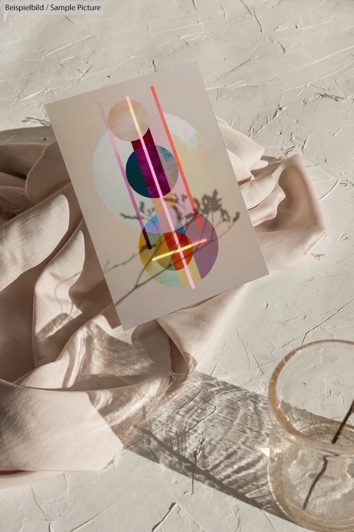 Abstract geometric art on paper with colorful circles against textured surface and draped fabric; shadow cast in foreground.