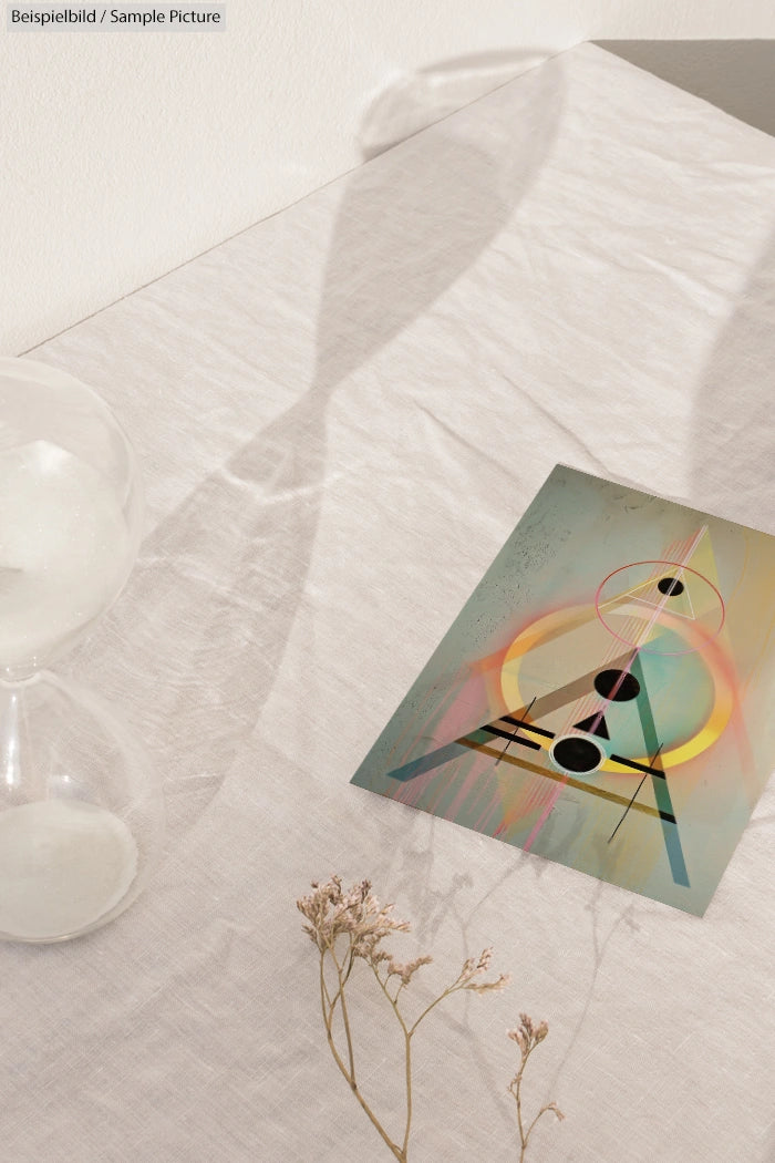 Abstract geometric artwork on a table with an hourglass and dried flowers casting a shadow.